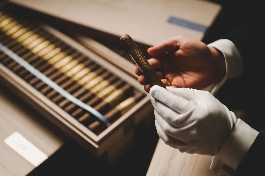 Hand with a white glove holds a cigar, cigar box in the background